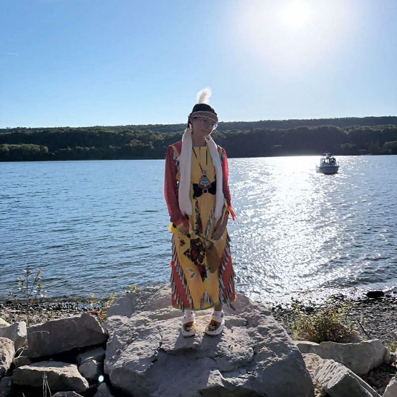 Image taken by Richard Panton of an indigenous woman standing on a large rock by the shore of a lake.